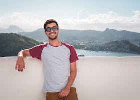 Young man in sunglasses leaning on a balcony with a scenic city view in the background.