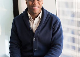 A professional black man smiling while seated by a window, embodying modern business vibes.