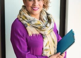 Smiling woman in a purple dress and scarf holding a tablet indoors.